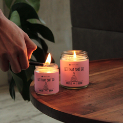 Person lighting a pink candle labeled 'Let That Shit Go' on a wooden surface with a plant in the background.