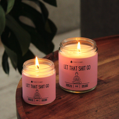 Two pink candles labeled 'Let That Shit Go' on a wooden surface with a plant in the background.
