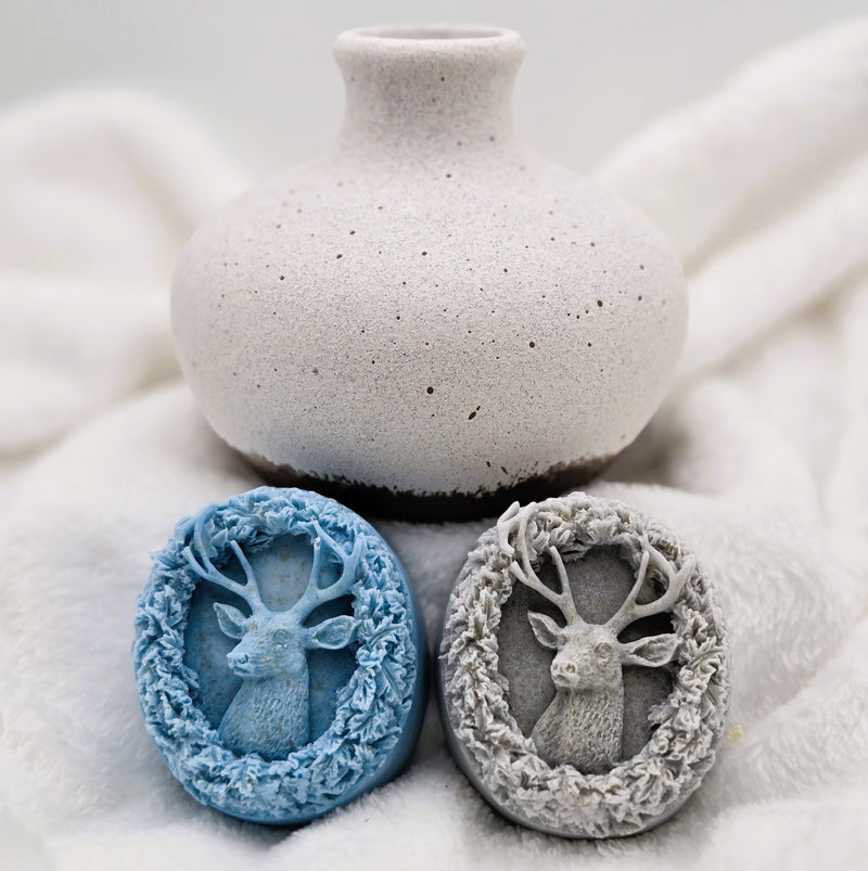 Two deer-shaped soap bars with a textured vase in the background on a soft surface.