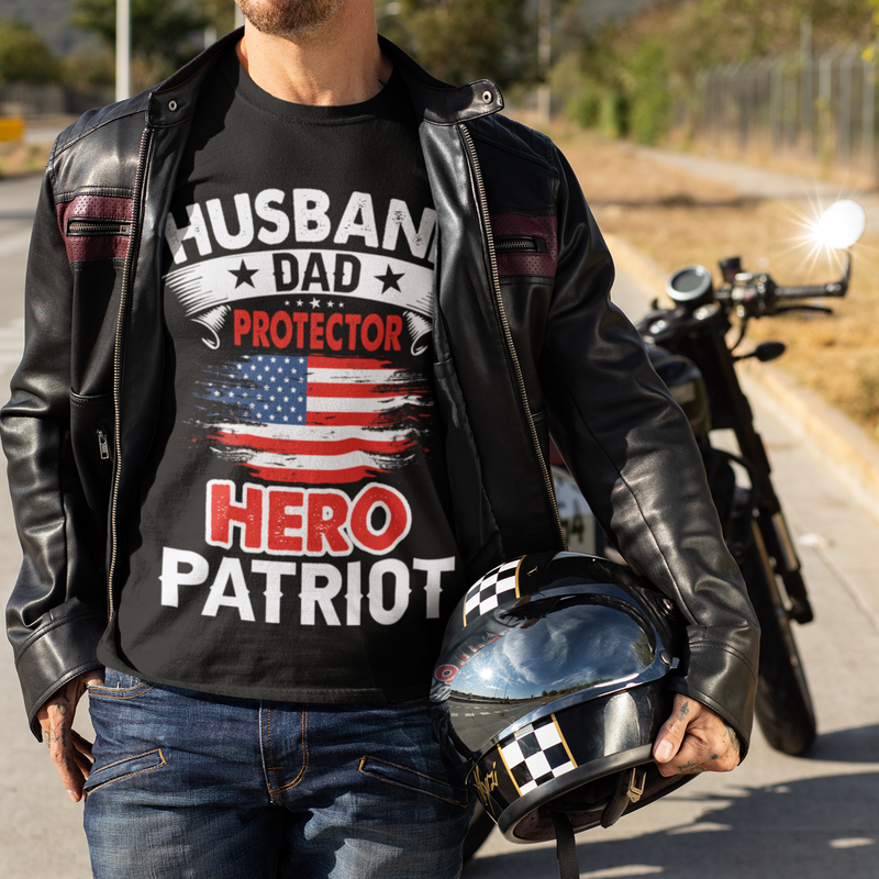 Man wearing a black t-shirt with 'Husband Dad Protector Hero Patriot' text and an American flag, holding a helmet on a road with a motorcycle.