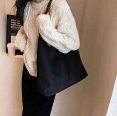 Person wearing a white knitted sweater and black pants against a beige wall holding a bag.