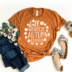 Orange t-shirt with 'fall breeze autumn leaves' text, surrounded by jeans, shoes, and decorative items on a white background