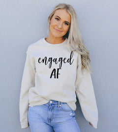 Woman wearing a sand color sweatshirt with 'engaged AF' text against a gray background