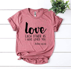 Pink t-shirt with 'love each other as I have loved you' text on a white wooden surface with a plant.