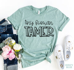 Dusty blue t-shirt with 'tiny human Tamer' text, paired with jeans, flowers, and sneakers on a white background.