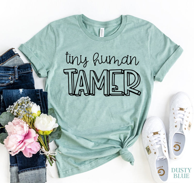 Dusty blue t-shirt with 'tiny human Tamer' text, paired with jeans, flowers, and sneakers on a white background.