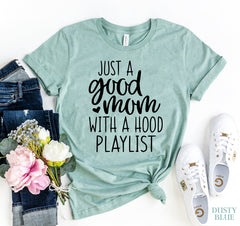 Dusty blue green t-shirt with 'Just a good mom with a hood playlist' text, paired with jeans, sneakers, and flowers on a white background.
