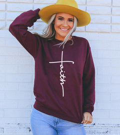 Woman wearing a maroon sweatshirt with 'Faith' and a cross, yellow hat, and blue jeans against a light brick wall.