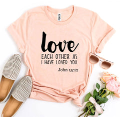 Peach t-shirt with 'love each other as I have loved you' text, paired with flats, sunglasses, and flowers on a white background.