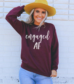 Woman wearing a maroon sweatshirt with 'engaged AF' text, yellow hat, and blue jeans against a light brick wall.