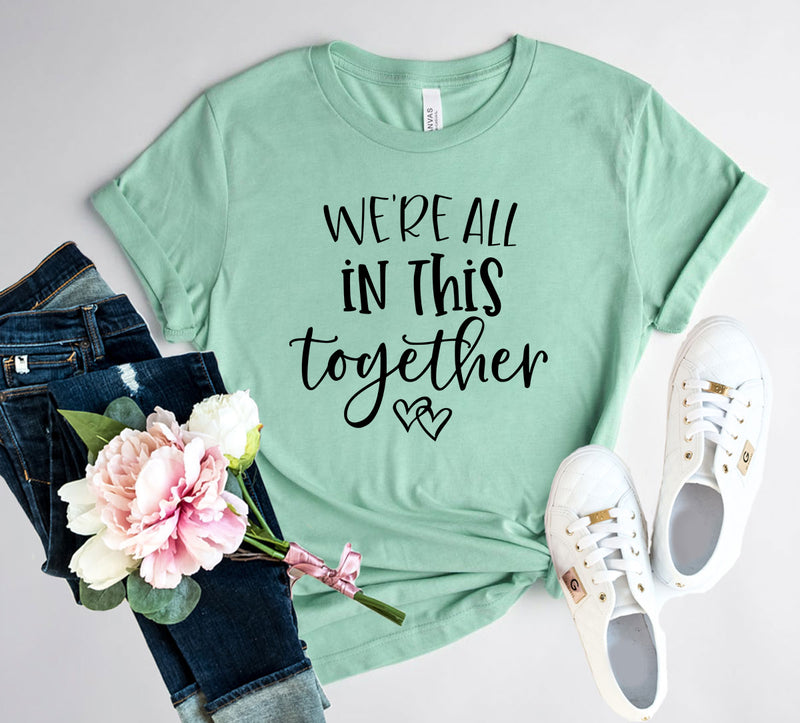 Mint green t-shirt with 'We're all in this together' text, paired with jeans and sneakers on a light gray background.