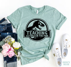 Heather dusty blue t-shirt with dinosaur graphic and 'Teaching is a walk in the park' text, paired with jeans, flowers, and sneakers on a white background.