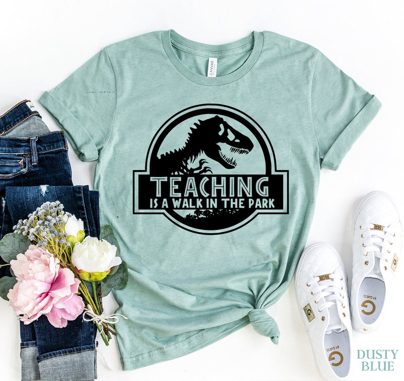Heather dusty blue t-shirt with dinosaur graphic and 'Teaching is a walk in the park' text, paired with jeans, flowers, and sneakers on a white background.