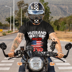 Person riding a motorcycle wearing a black t-shirt with text and graphics, on a road with trees and mountains in the background.