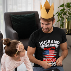 Man wearing a 'Husband Dad Protector Hero' t-shirt wearing paper crown indoors looking at a child.