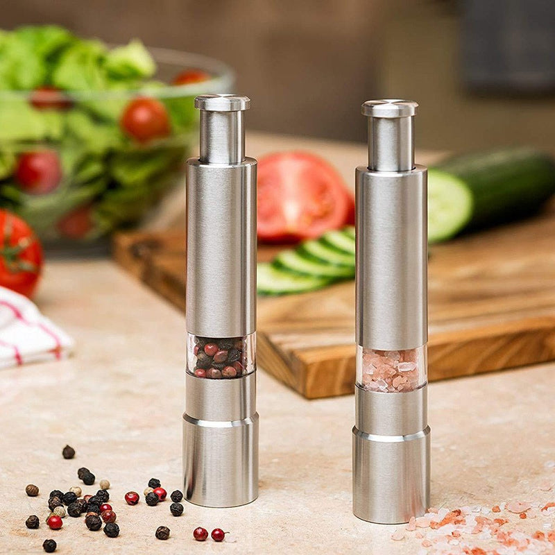 Two silver pepper mills on a kitchen counter with salt and pepper visible, surrounded by fresh vegetables.