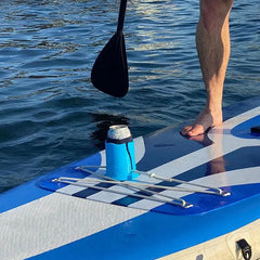 Person on a paddleboard with a blue and white board, holding a paddle, and a can cooler with a can on a checkered mat.